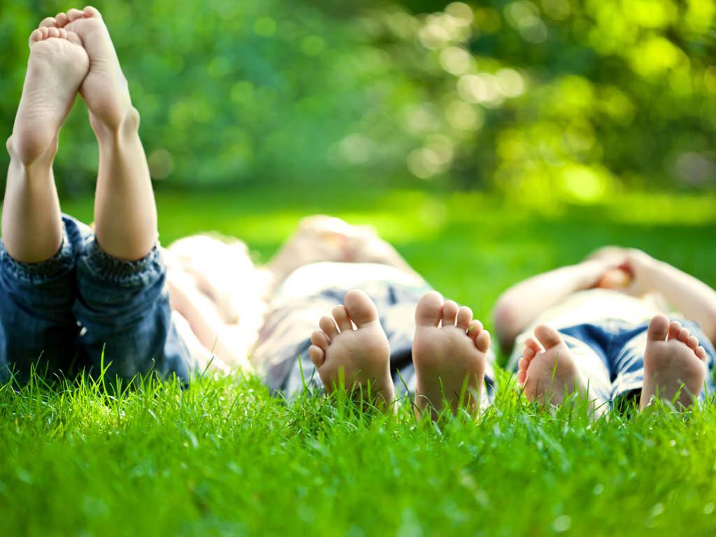 Three young people lay side by side in a grassy area. The photo is taken from the ground, viewing their feet. The person on the left has their feet raised into the air. A line of trees is in the background, out of focus.