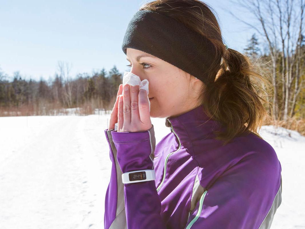 A woman stands outside in winter, wearing workout clothes including a jacket and headband. She holds a tissue up to her nose. Snow is on the ground and trees are in the background.