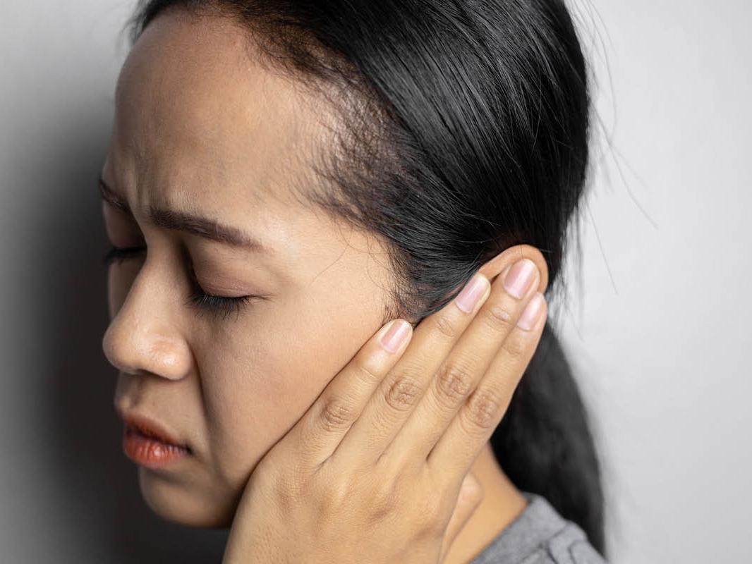 A close-up of a woman with her eyes closed and an irritated expression on her face. She is covering her left ear with her left hand.