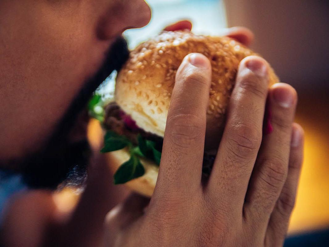 A close-up of a man with a short beard and mustache holding a burger on a bun, about to take a bite out of it. He holds the burger with both hands.
