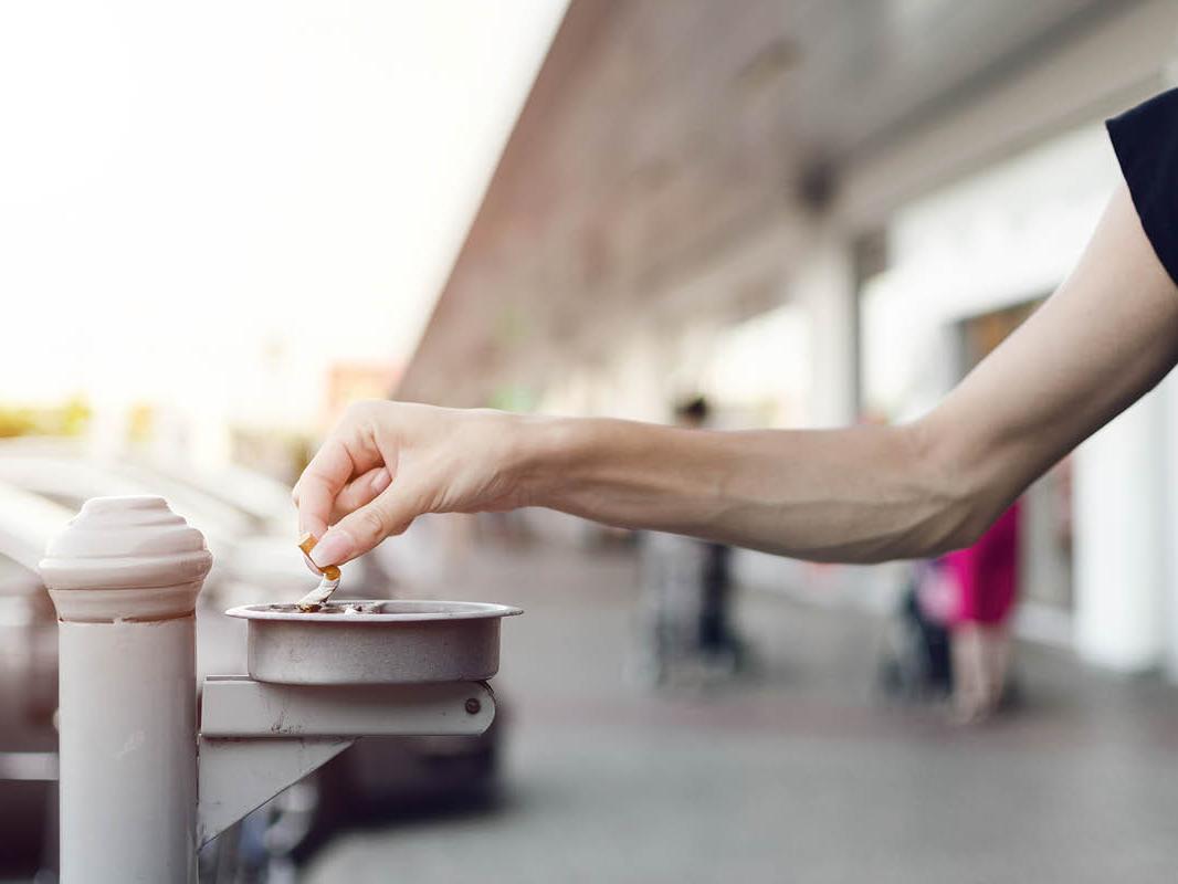 An outstretched hand holds a cigarette butt as its remnants are snuffed in an ashtray, which is mounted to a pole outdoors. Vehicles and a building are in the background, slightly out of focus.