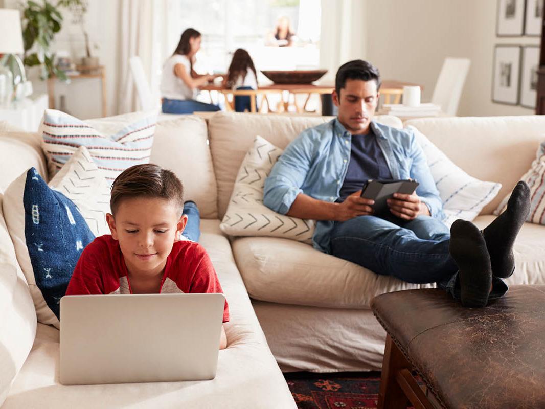 A man and young boy each use electronic devices while on a couch. A woman and young girl are in the background, slightly out of focus, sitting at a table using devices.