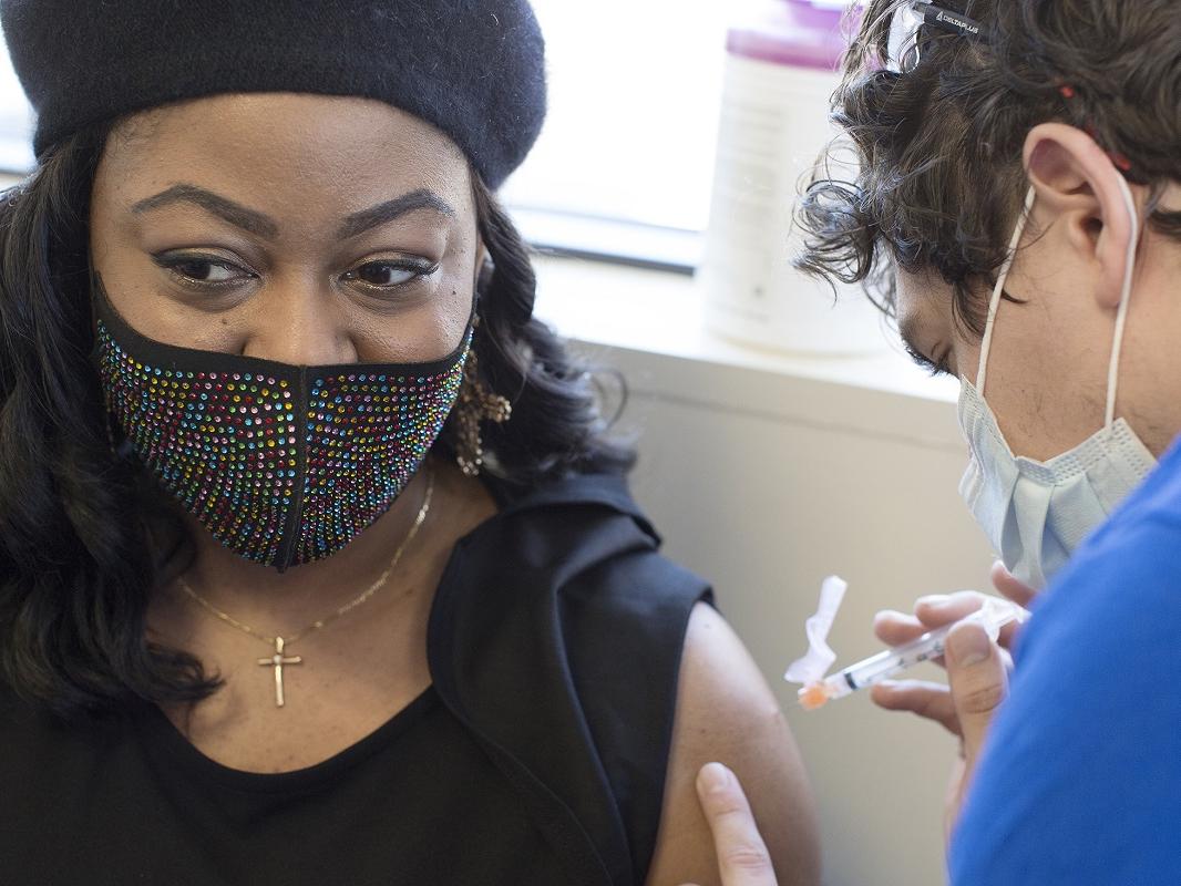 Tamika Washington, who wears a jeweled face mask, a cross necklace and a beret, looks away from her bare arm as a health care worker prepares to insert a syringe filled with the COVID-19 vaccine.