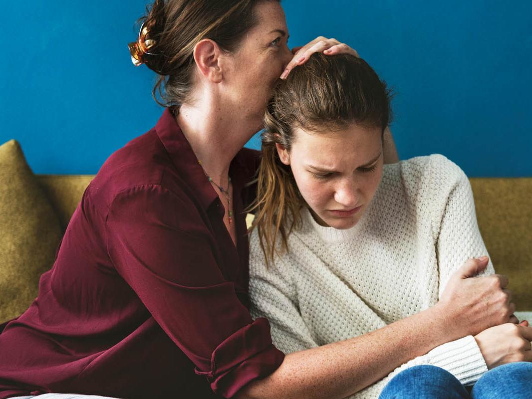 A woman sits next to a teenage girl on a couch. The woman has her right arm around the girl and her left hand on her head, and her face resting on the girl’s head.
