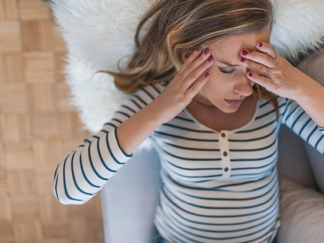 A woman lays on a couch with her head on a pillow. Her eyes are closed and she has both hands placed on her forehead.