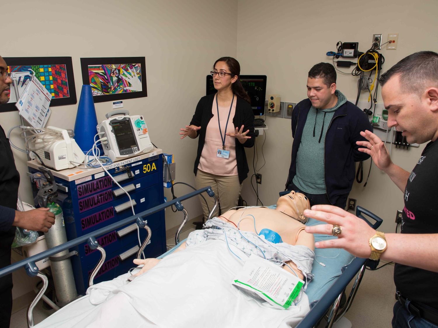 Dr. Lidys Rivera Galvis and students stand around a mannequin that lays on a wheeled-stretcher and is connected to heart machine.