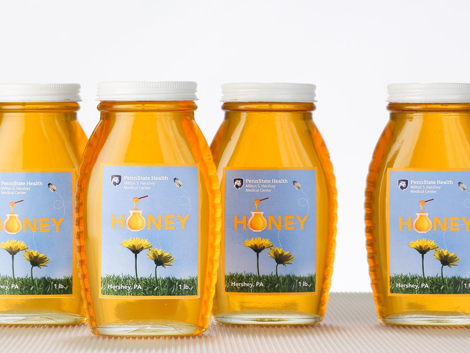Four jars of honey bear labels that have the Penn State Health logo and an image of two dandelions and the word "Honey" in yellow letters. The background of the label is blue.