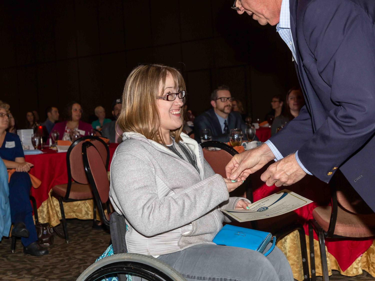Megan Patrick, wearing a sweatshirt and jeans, smiles at a man in a sport coat who is placing something in her hand. In her other hand, she holds a certificate.