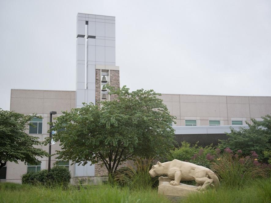 A statue of the Penn State Nittany Lion at the Penn State Health St. Joseph campus appears in profile in front of a row of trees and weeds. Behind the scene rises a glass tower and bells and windows.