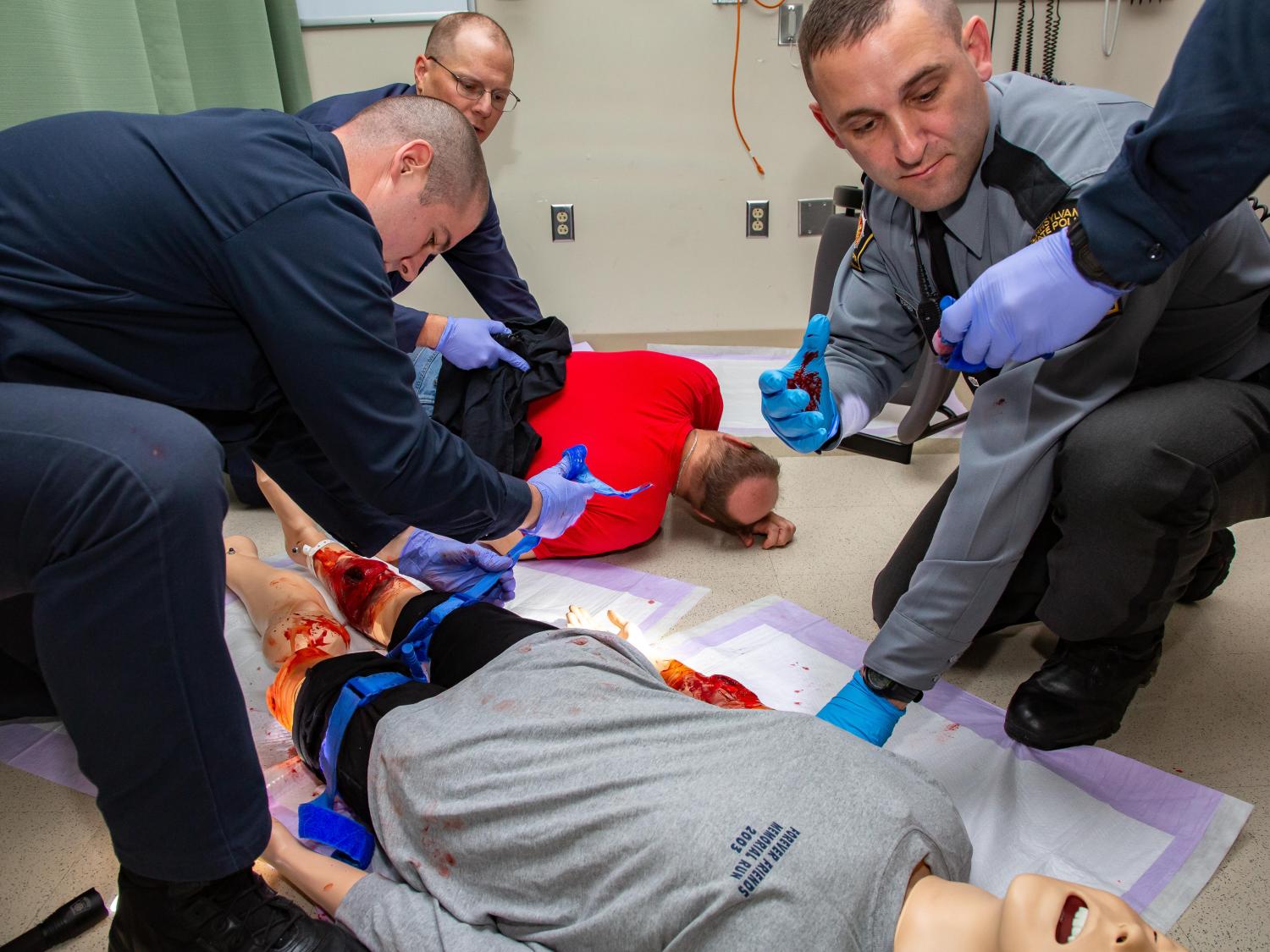 Three Pennsylvania State Police cadets learn how to apply a tourniquet during Stop the Bleed training. A man in the front-left tightens a tourniquet strap on a dummy, which has a fake gunshot wound on its right leg and blood on its left leg. A man in the back-left holds a cloth to the back of an actor. A man in the front-left reaches for a device with his gloved hand, which is covered in fake blood. The arm of another cadet reaches in holding a device. They are working in a clinical room.