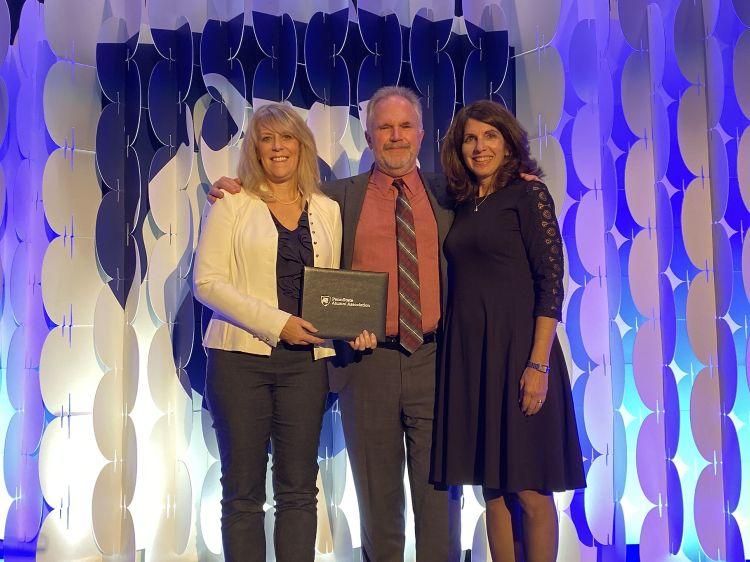 Two women stand on either side of a man as they hold a Penn State Alumni Association award.