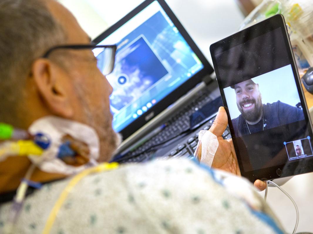 A man in a hospital gown in his hospital bed holds up an iPad on which a man is smiling back at him. A laptop computer is in the background, slightly out of focus.