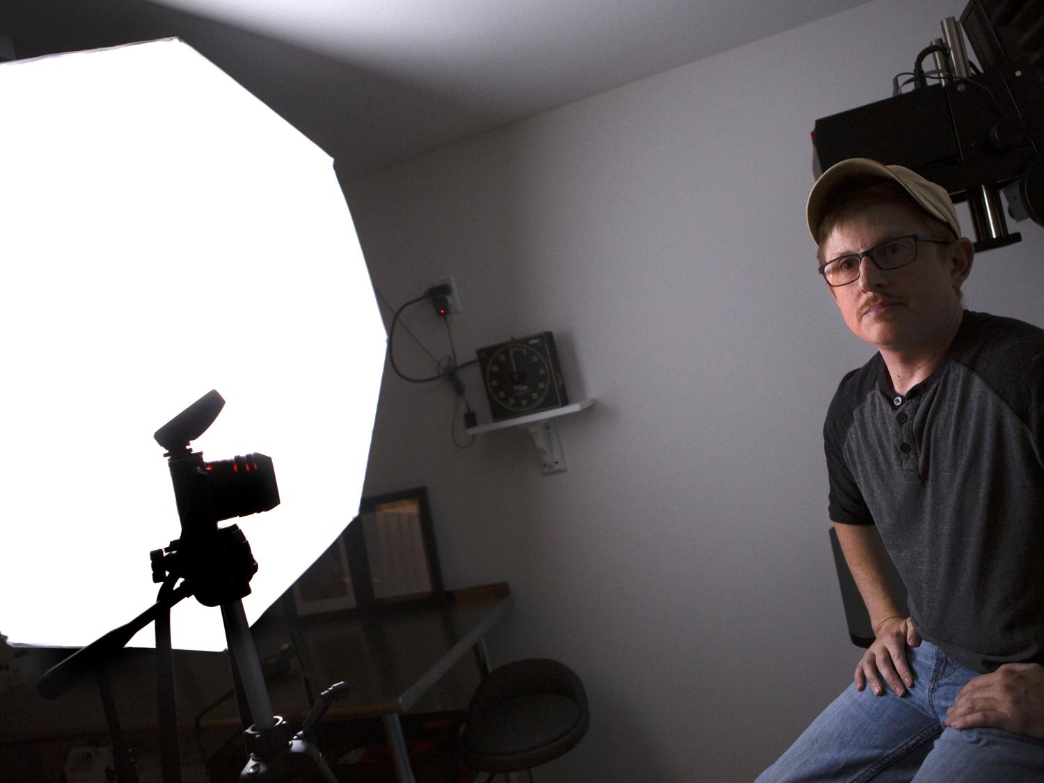 A man wearing a ballcap, shirt and jeans sits on a stool in a photo studio. A camera and light panel are on the left side of the photo.