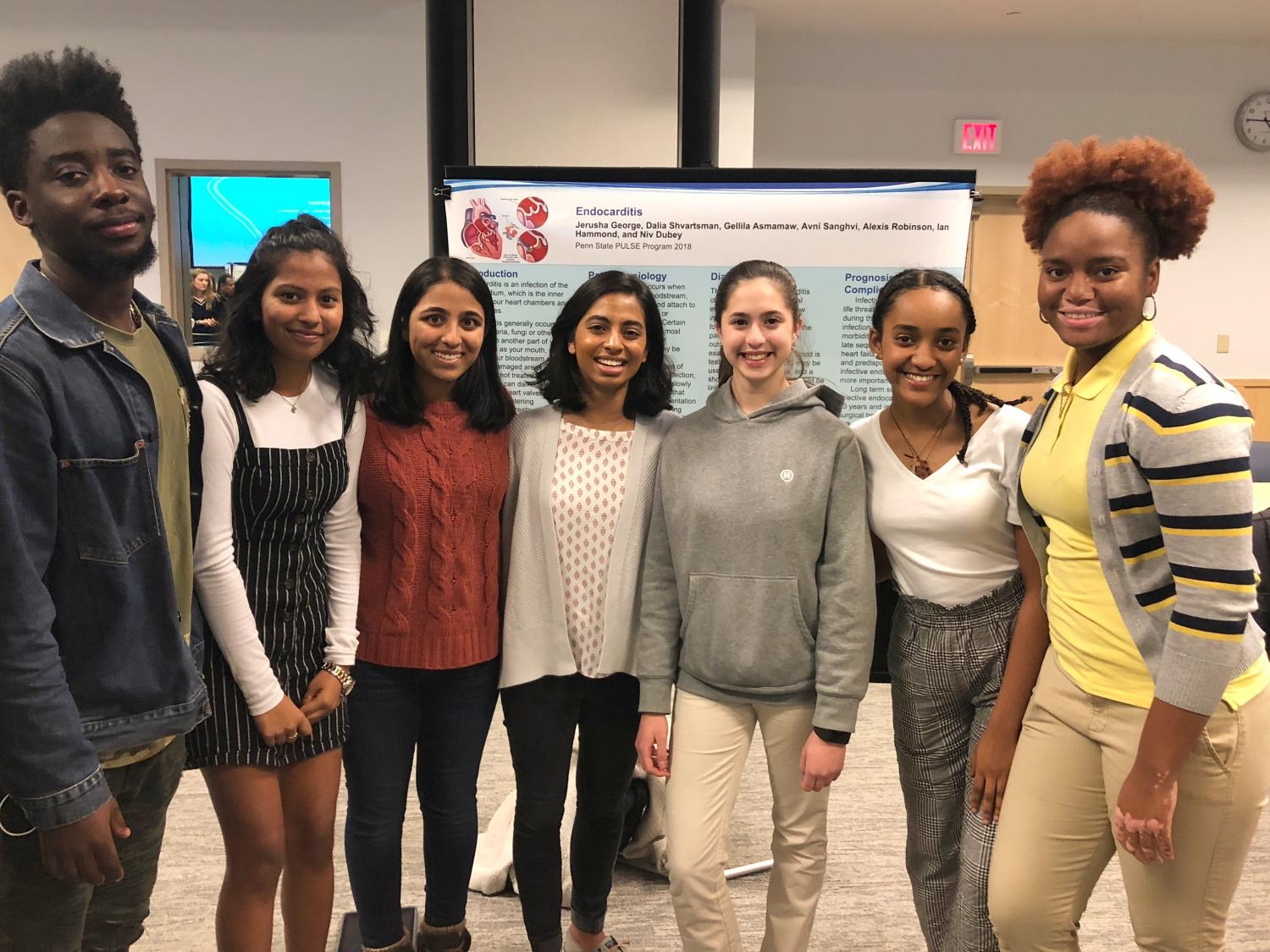 Seven high school students who won the 2018 poster session at Penn State College of Medicine’s PULSE program smile in front of a poster presentation on endocarditis. One young man and six young women stand in a row and smile.