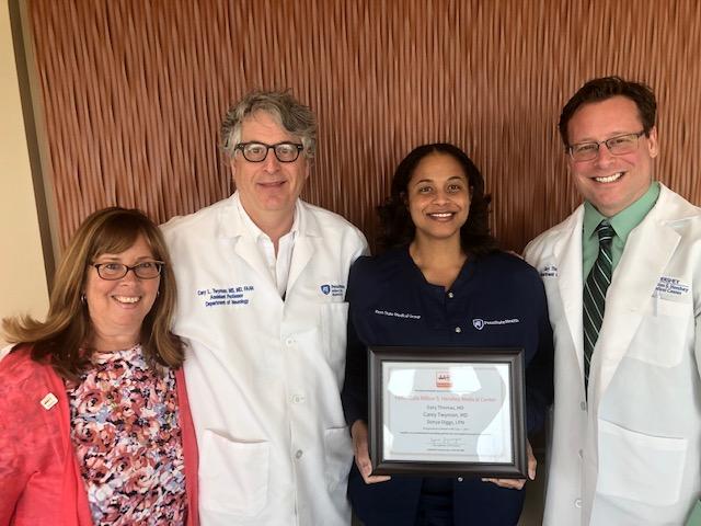 Two men and two women stand in a row, smiling, posing for a photo, against a brown textured backdrop. Three of them wear clothing with the Penn State Health logo. One woman holds a framed certificate.