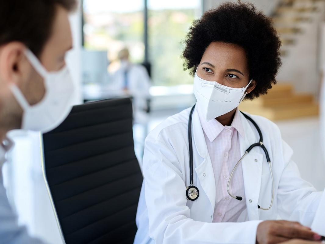 A Black woman doctor wearing a protective face mask talks to a male patient at a clinic. The doctor has a stethoscope around her neck and is sitting in a chair. The male patient is wearing a face mask and a button-down shirt.