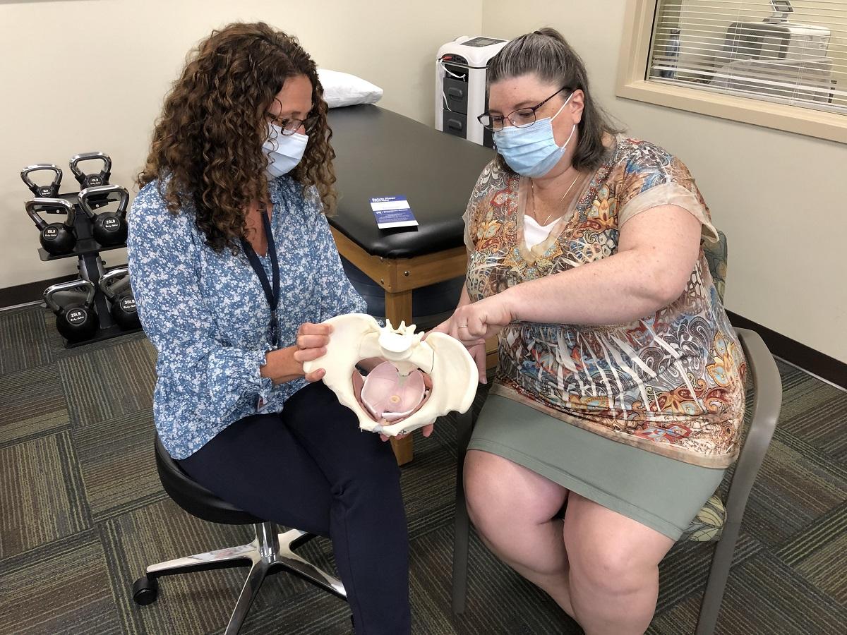Pelvic floor physical therapist Margaret Homan, left, holds a model pelvis in her hands as she discusses with Pamela Betz how therapy improves symptoms of pelvic floor dysfunction. They both are seated and wearing face masks.