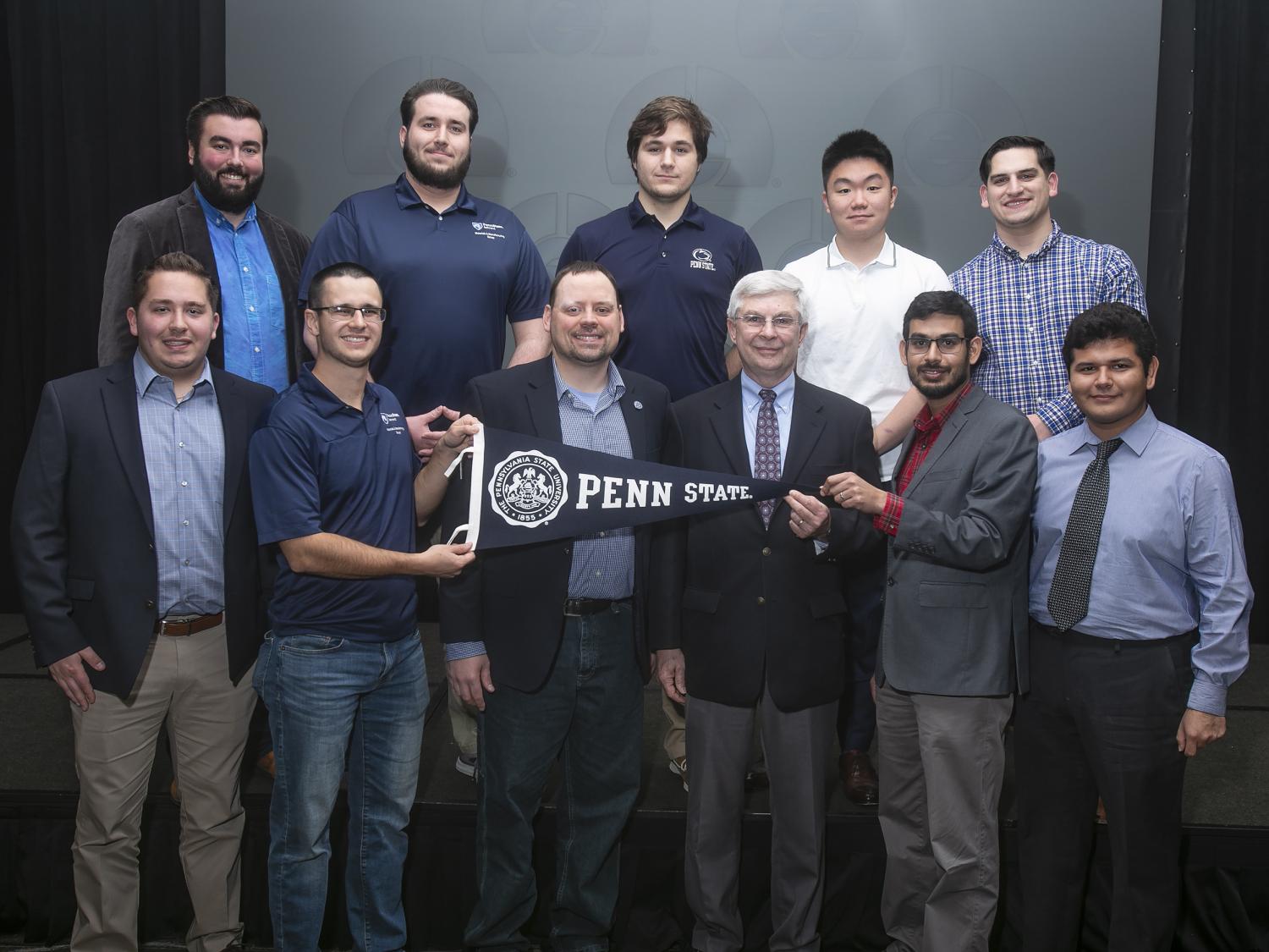 11 men stand together on a stage while two are holding up a Penn State pennant flag.
