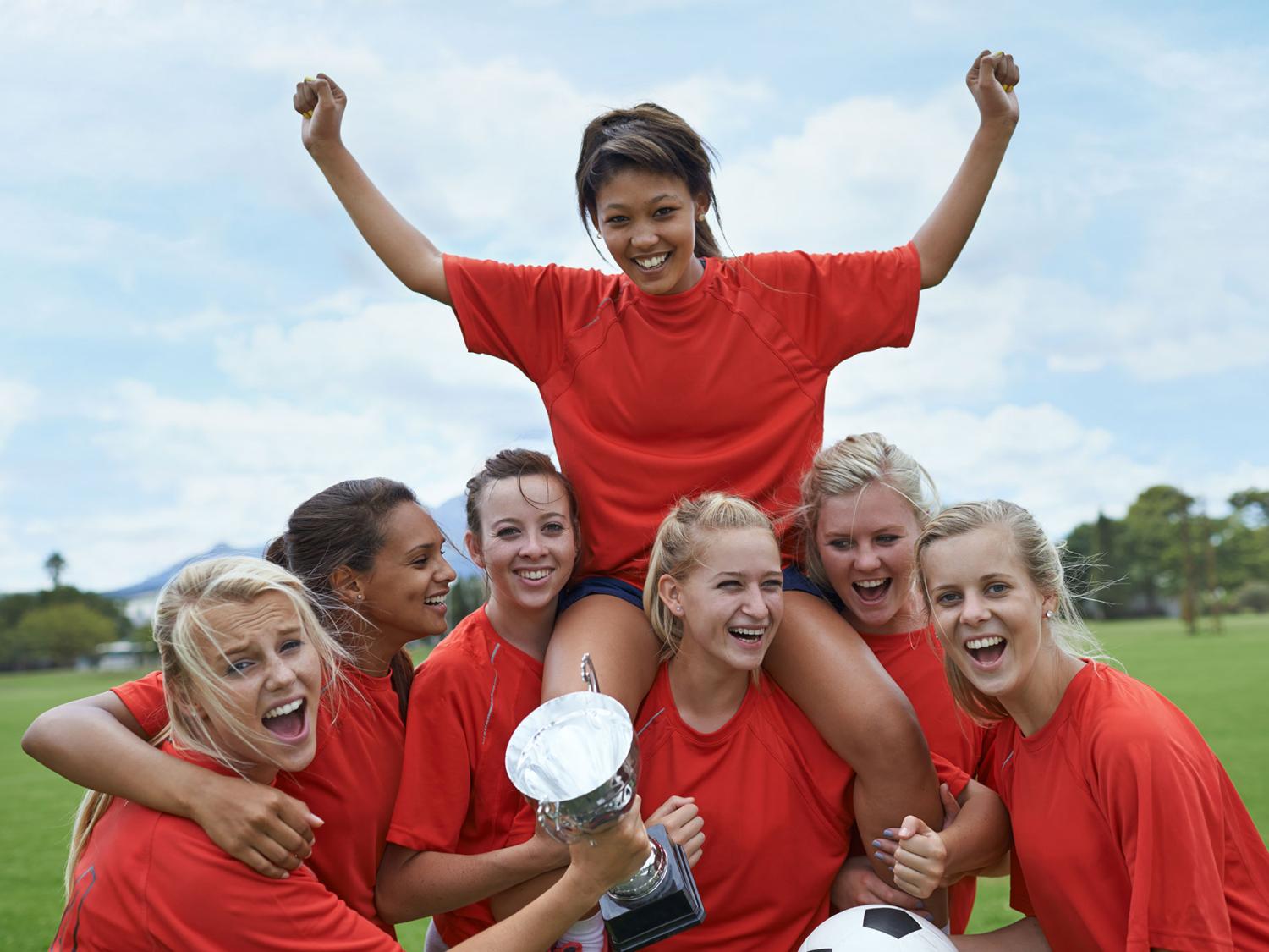Women's sports team in a huddle, holding a trophy and raising a teammate on their shoulders.