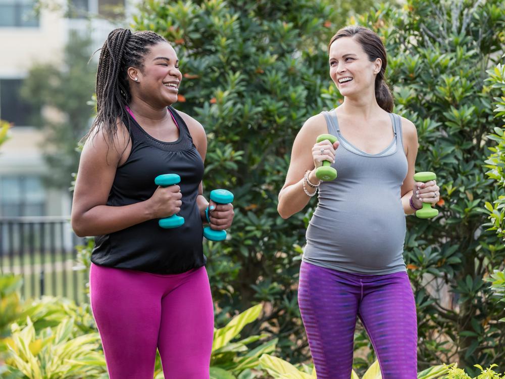 Two women in exercise clothing walk side-by-side, each holding small hand weights. They look at each other, smiling. Bushes are in the near background; farther in the background is an apartment building, slightly out of focus.