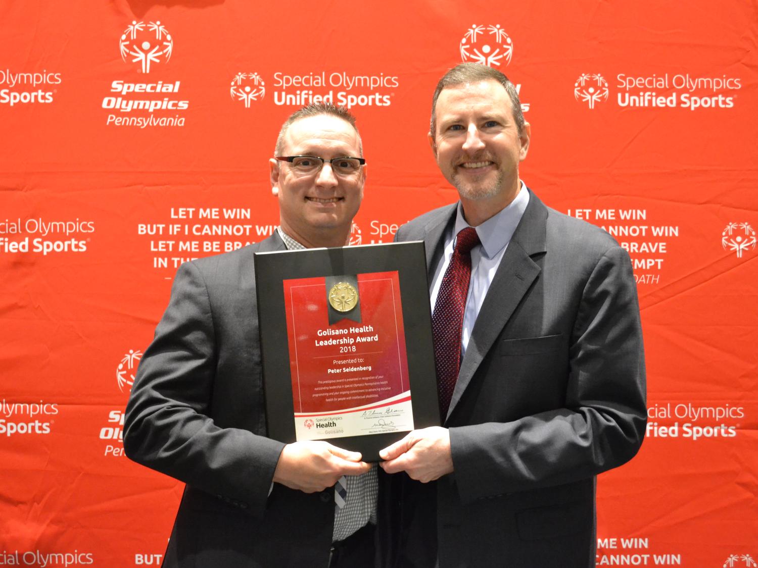 Two men in suits pose for a photo, each holding a corner on an award certificate. The backdrop bears the Special Olympics Unified Sports logo.