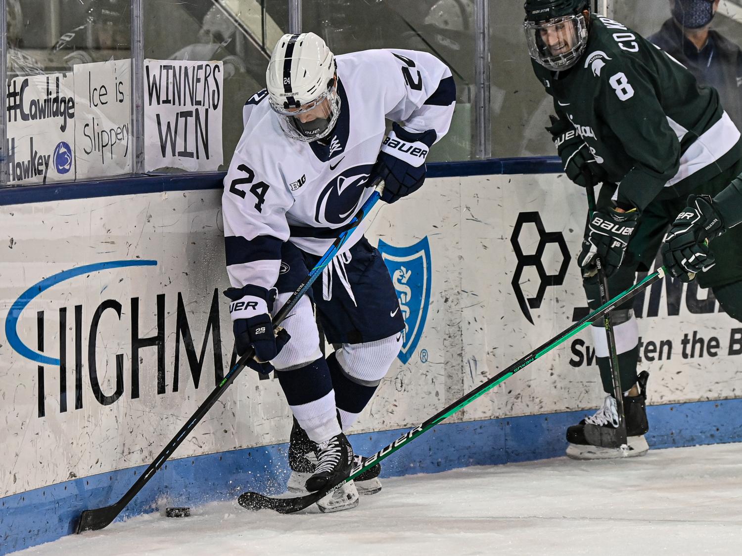 A hockey player dressed in navy blue and white protective gear and a helmet goes after a hockey puck along the edge of an ice rink. 