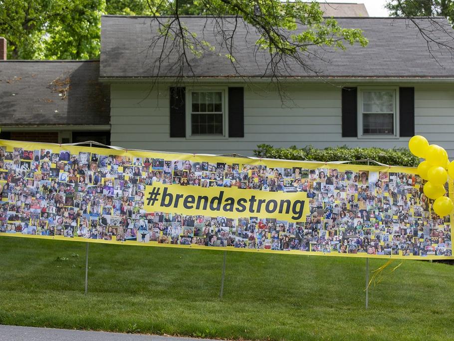 Two women stand at either end of a sign on wooden posts. They attach balloons to the edges of the sign, which sits on a lawn in front of a small house. The sign says “#brendastrong” and is awash in photos.