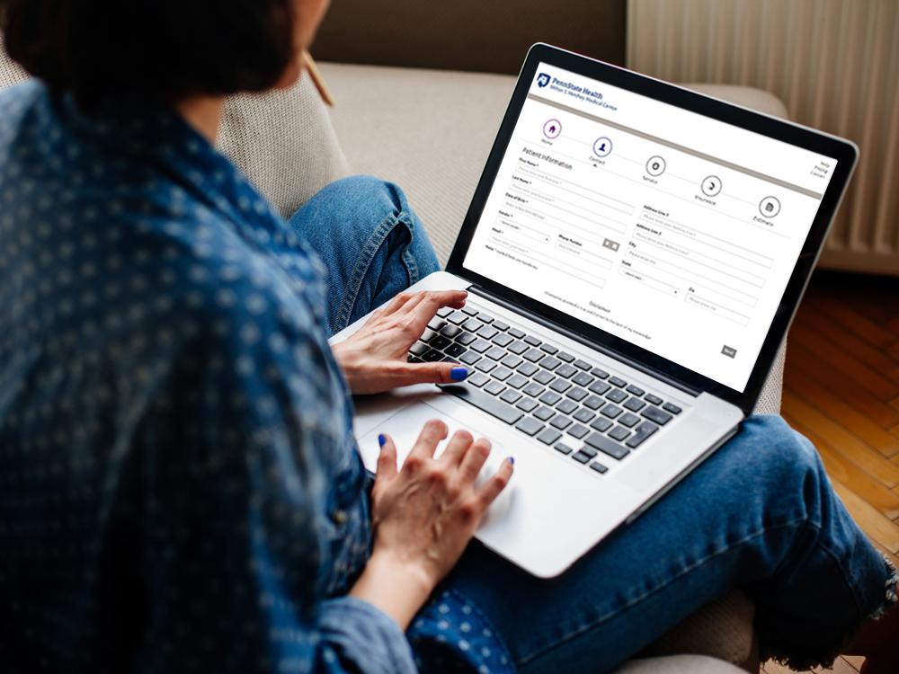 A woman sits on a couch, using a laptop. She wears a button-down shirt and jeans.