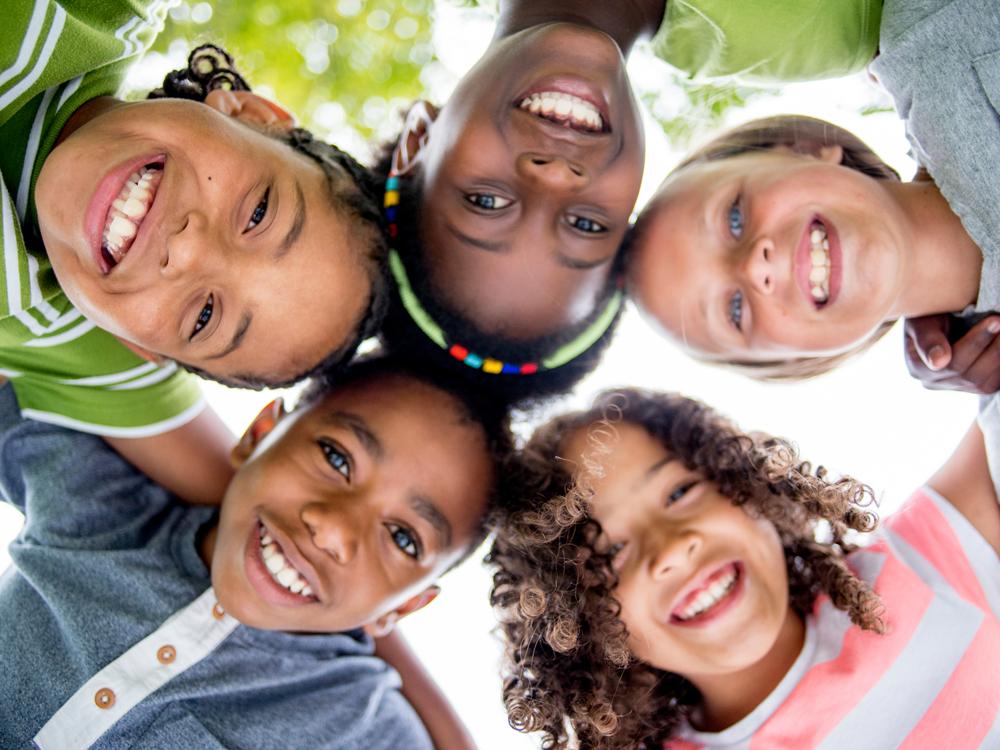 Five young children put their heads together, standing in a circle and looking downward at the camera. They are all smiling.
