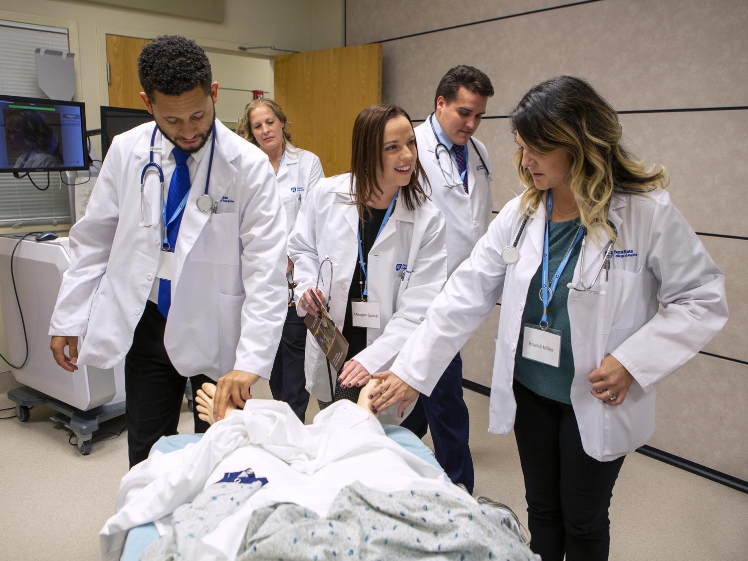 A man and two women touch the feet of a mannequin that is lying on a table. Behind them a woman and man stand. All five are wearing white lab coats. Behind them is a cart with two monitors and a door.