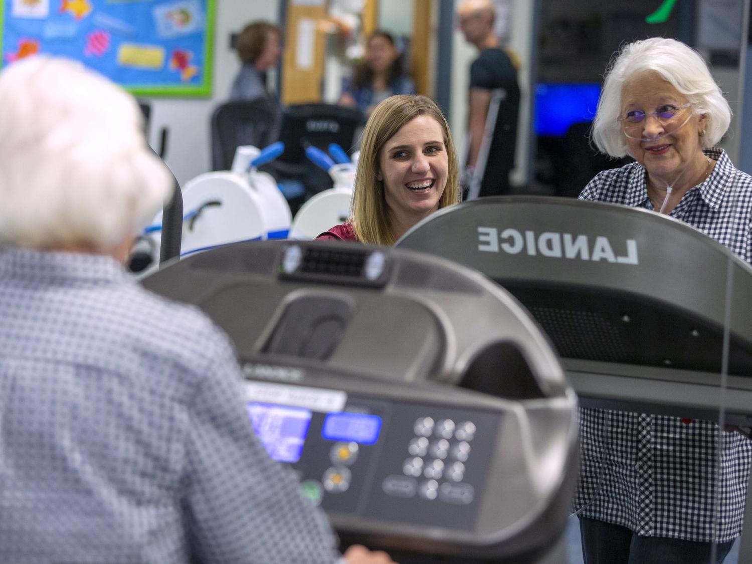 A young woman smiles as an older woman wearing a nasal cannula and a checkered dress and standing on a treadmill. They are in the Health and Wellness Center on the Hershey Medical Center campus. Three people are standing behind them out of focus.