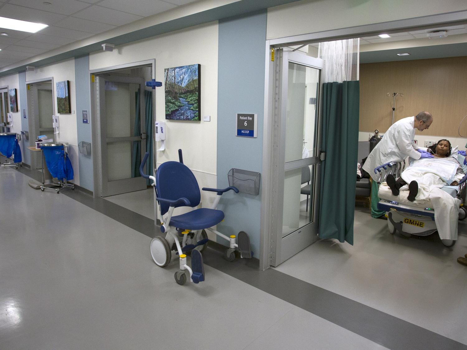 A physician and two other care providers treat a patient who is lying in a bed in Hershey Medical Center’s new Radiology Outpatient Care Unit. A long hallway with a tiled floor shows five rooms.