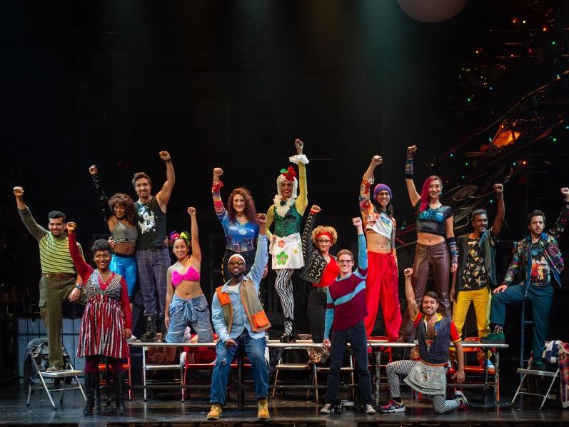 A diverse group of people stand on a stage and each raise a fist into the air.