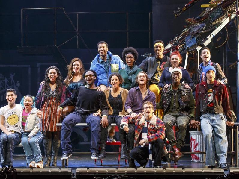 The cast of “Rent” sits on top of a table while singing a musical number.