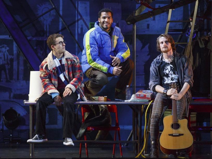 Three characters from the musical “Rent” sit on and stand near a table.