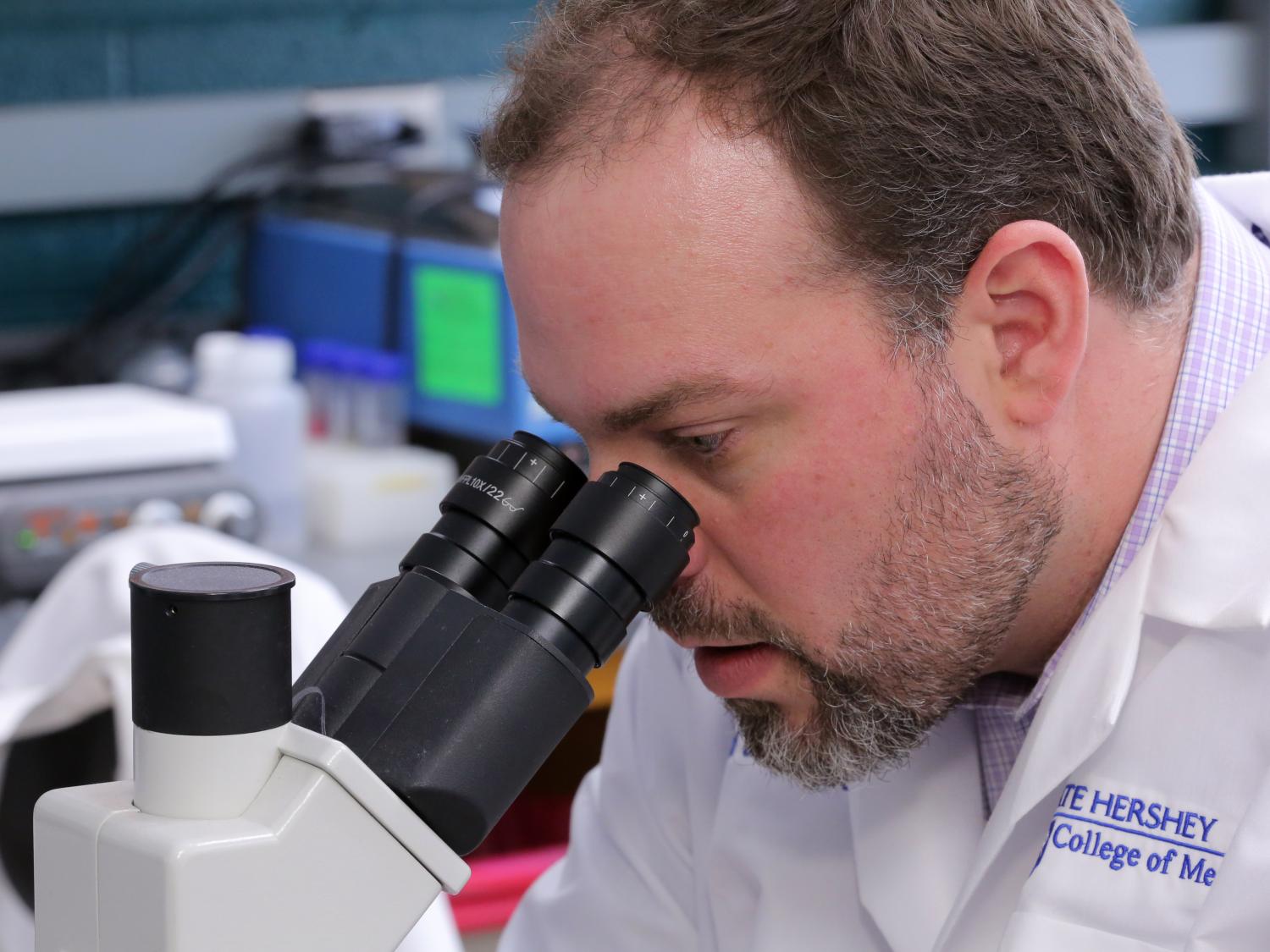 A close-up of Dr. David DeGraff, in a white lab coat, looking into a microscope.