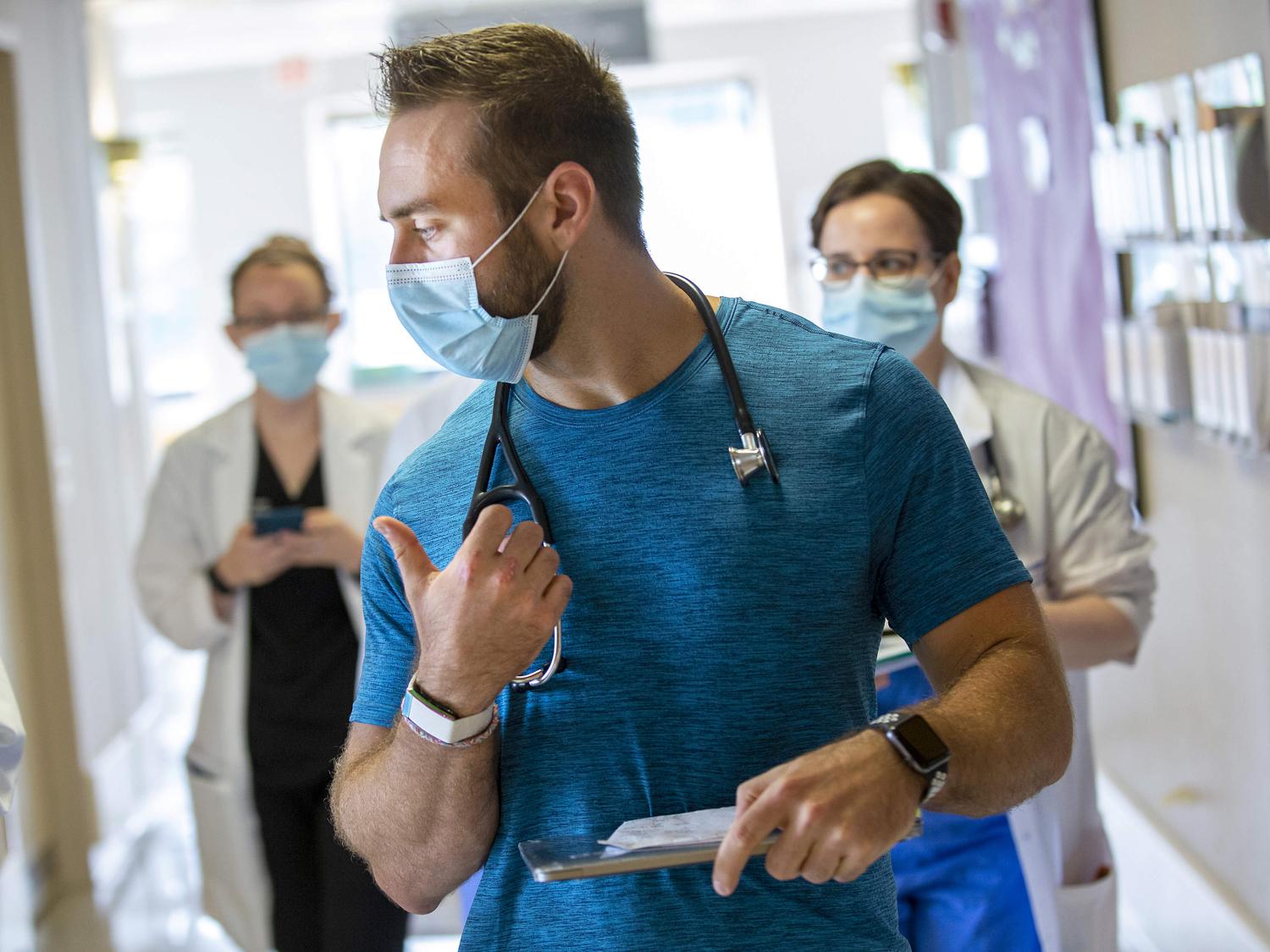 Dr. Alexander Hajduczok, a third-year internal medicine resident at Penn State Health Milton S. Hershey Medical Center, sports a wearable device manufactured by WHOOP on his right wrist. Two other physicians are behind him, walking down a hallway.