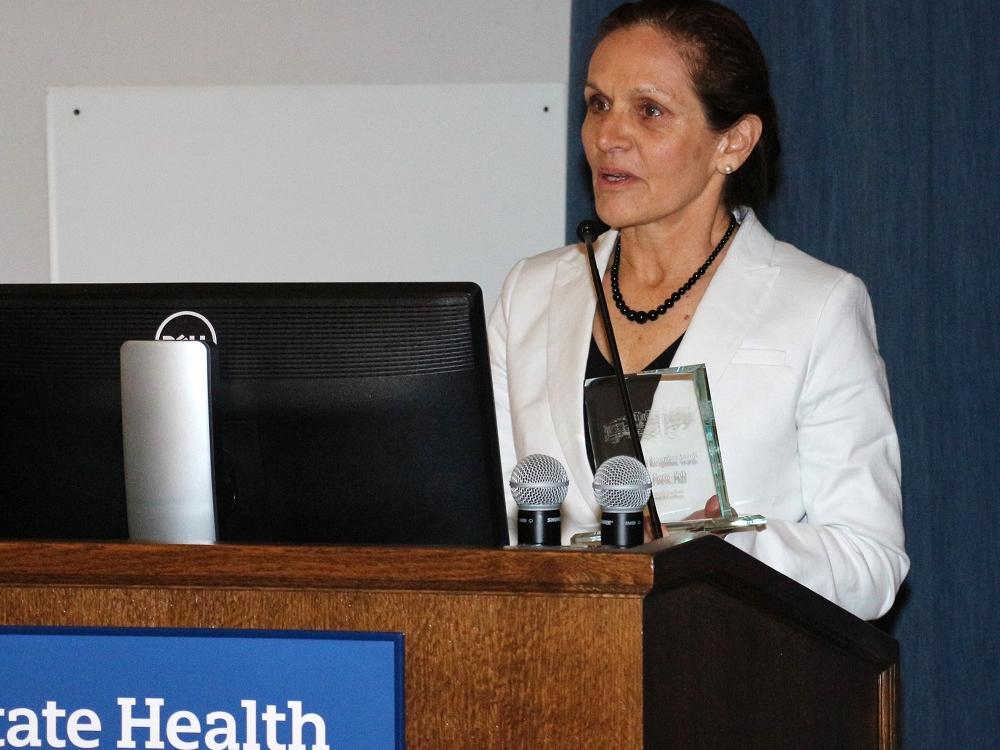 Joanna Floros, wearing a white coat and holding a glass award, speaks at a lectern.