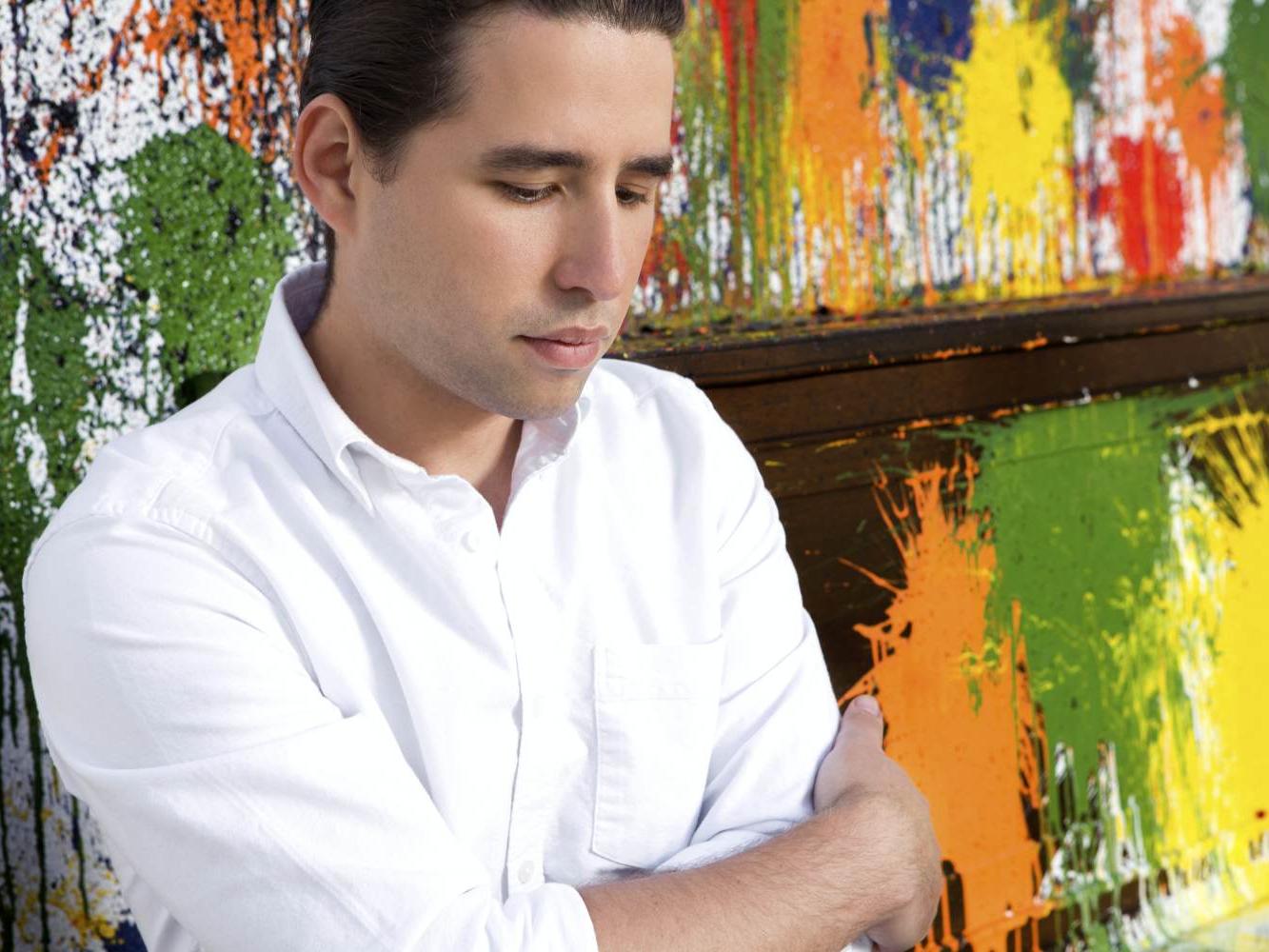 Cuban pianist Alfredo Rodríguez stands with his arms crossed in front of a piano covered with splattered paint.