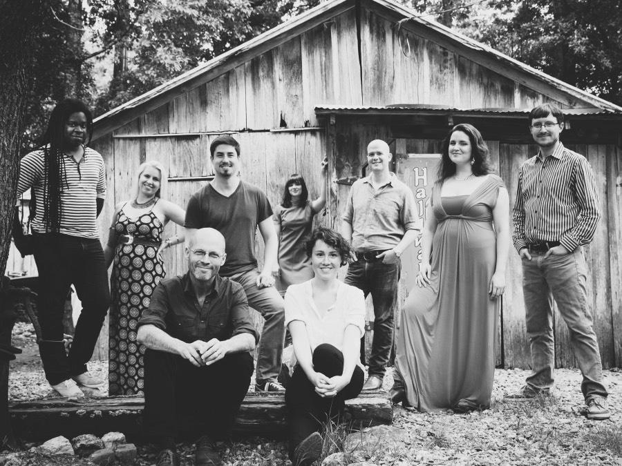 Seven singers stand in front of a barn while Roomful of Teeth Artistic Director Brad Wells and eighth Roomful of Teeth vocalist sit on a log.