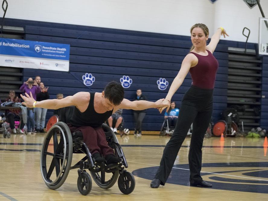 A man and a woman in leotards raise the arms on a gymnasium floor. The man in a wheelchair, bows at the waist. Behind them several people snap photos on cell phones or applaud.