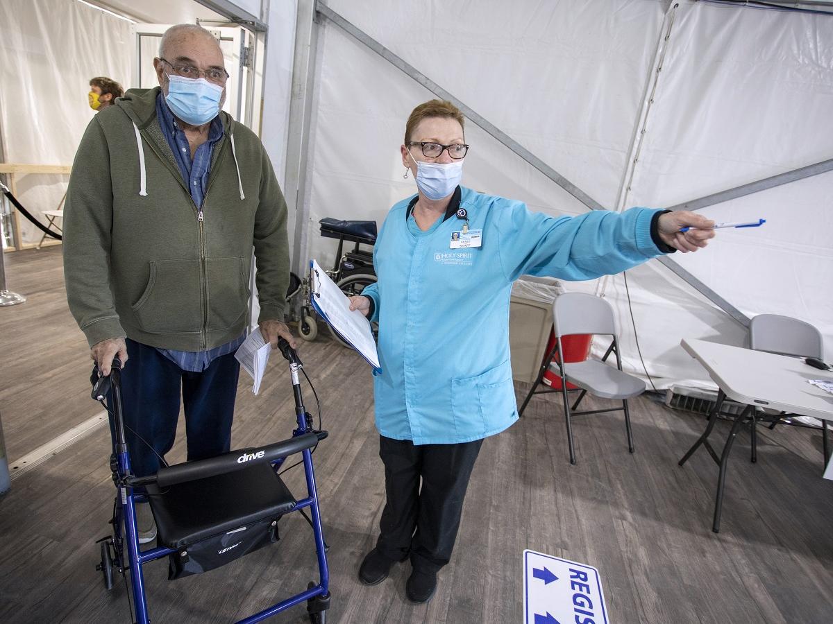 Renee still points with her left arm to direct Lee Jackson, who stands next to her and uses a walker, to a spot across the COVID vaccination tent.