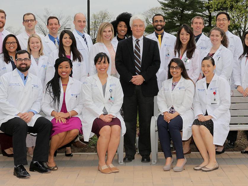 Dr. John Repke, wearing a suit, poses outdoors with 26 residents - some seated, some standing -- all of whom wear white clinical coats.