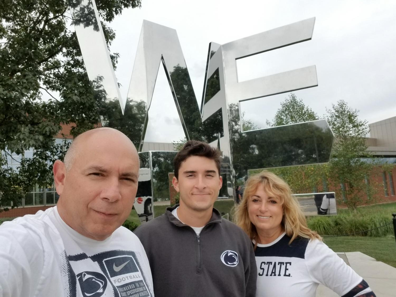 Three people look and pose for a camera in front of the We Are statue on Penn State's University Park campus. 