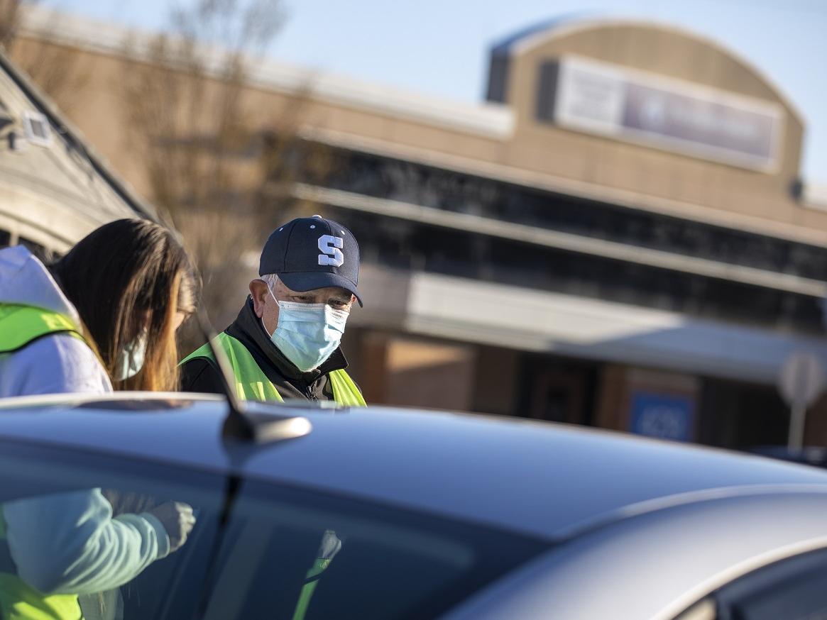 A man in a ball cap with an S on the front and a surgical mask stands next to a woman in a mask and a hoodie, partially obscured by a car.