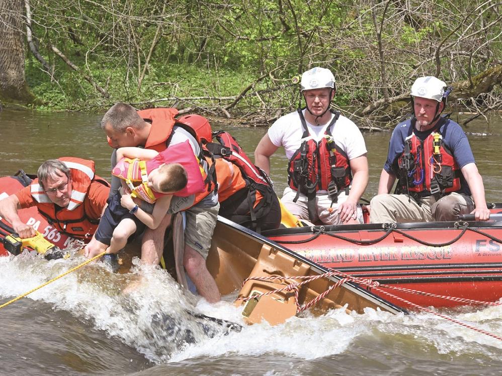 Rex Carmichael, wearing glasses and a life vest, saws tree roots to free a child in a creek as another paramedic holds onto the boy, who is slumped over his arm. Three other paramedics, wearing helmets and life jackets, sit in a river rescue boat.