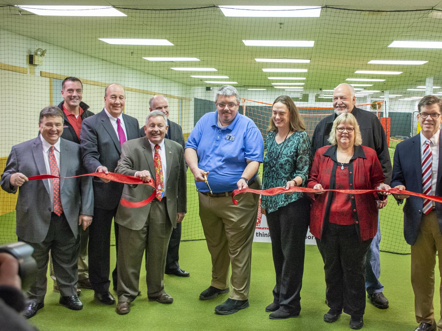 Several people stand on turf grass in a line while holding a red ribbon. Joe Medica stands in the center holding scissors to cut the ribbon.