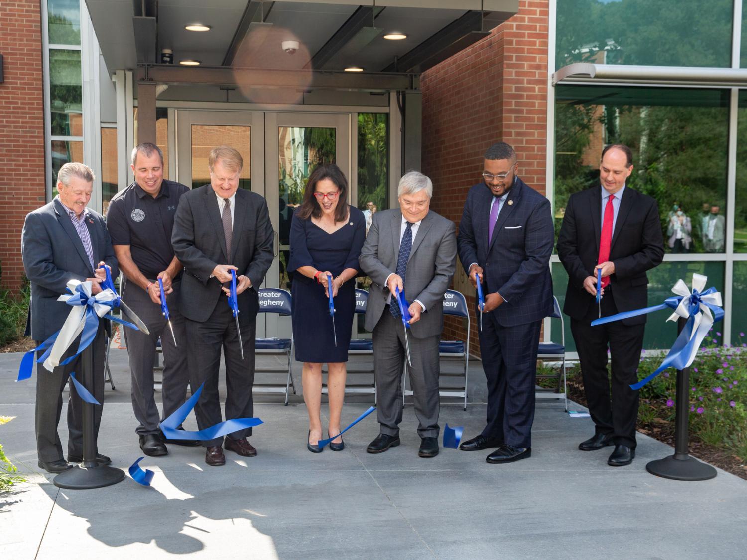 Group of people holding oversized scissors cut ribbon. 