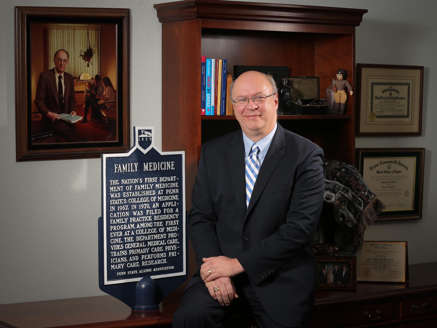 Dr. Mack Ruffin IV sits on the edge of a desk with a bookshelf in the background. A portrait appears on the wall, and Dr. Ruffin sits next to a plaque that reads "family medicine."
