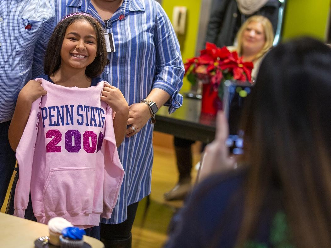 Ruth Vega smiles as she holds a sweatshirt up in front of herself that says Penn State 2000th. Her parents stand behind her. A poinsettia plant sits on a table in the background, with people sitting around it. 